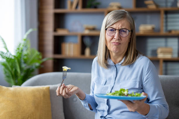 istock Woman with glasses sitting on sofa holding salad plate and fork with confused expression, surrounded by modern home decor 2180558082