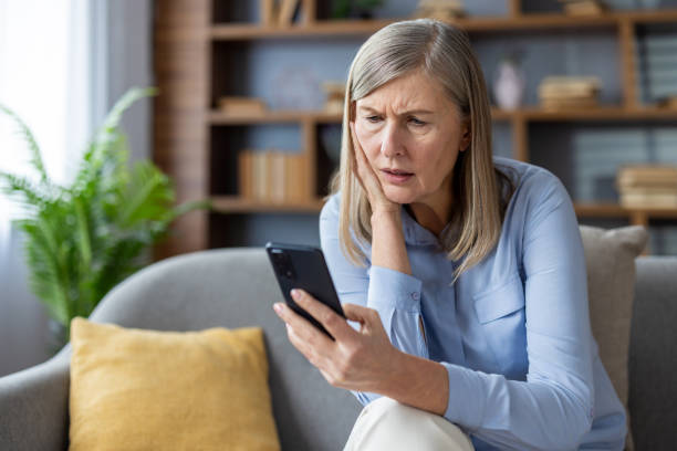istock Worried woman in blue shirt sitting on sofa while looking at smartphone, experiencing difficulty or confusion with technology at home. Mature lady holding phone with thoughtful expression. 2180558304