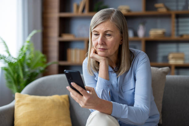 istock Mature woman using smartphone while sitting on a comfortable sofa in a modern living room setting, showcasing thoughtful expression as she engages with digital content on her phone. 2180558389