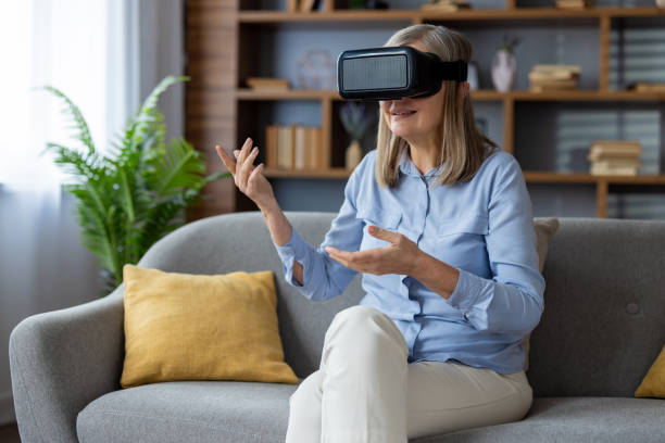istock Senior woman enjoying virtual reality experience at home, seated on a comfortable grey sofa wearing a VR headset, engaging with immersive technology in a relaxed environment 2180558544