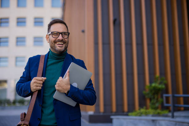 istock handsome and attractive man is returning from work, holding his laptop in his hands after a successful day full of challenges. Satisfied and happy, he walks down the street beside a modern building, enjoying every moment. 2180558905