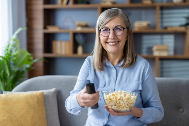 istock Smiling woman holding a bowl of popcorn and remote control while sitting on a cozy sofa. Enjoying leisure time at home with entertainment. Relaxed elderly lady ready to watch TV. 2180559491