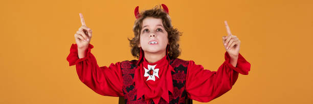 Panoramic view of cute boy in demon or vampire fancy outfit, with fangs and horns pointing and looking upwards in isolation