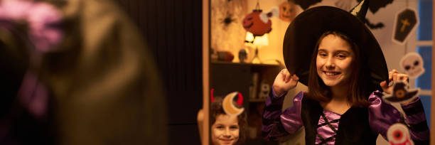 Banner of cute cheerful girl in costume of Halloween witch standing in front of mirror while getting ready for party against demon boy
