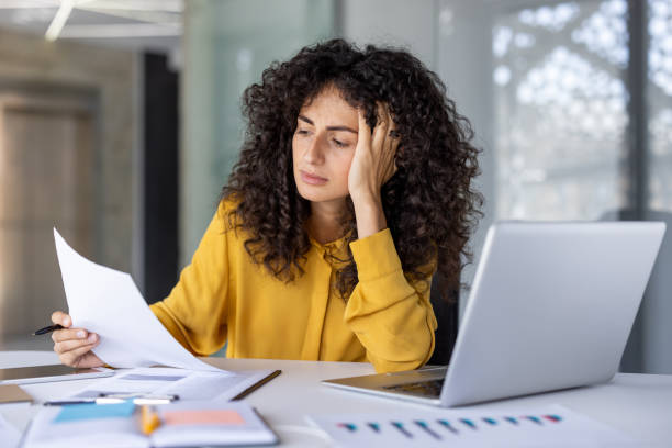 istock Hispanic businesswoman feeling stressed while working at laptop in modern office, analyzing documents, and multitasking 2180564751