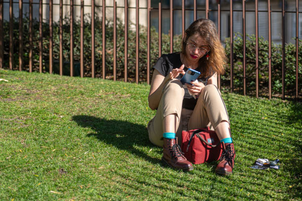 Porto, Portugal - September 13, 2024 : A woman sits on the grass, absorbed in her smartphone and enjoying the weather.
