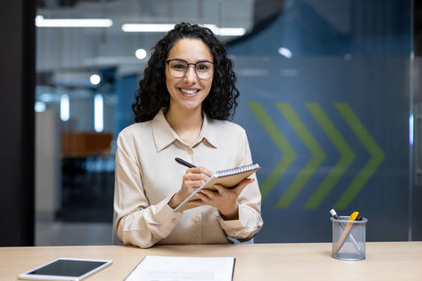 istock Smiling businesswoman holding notebook in modern office setting, appearing confident and professional while looking at camera during work breaks 2180572709