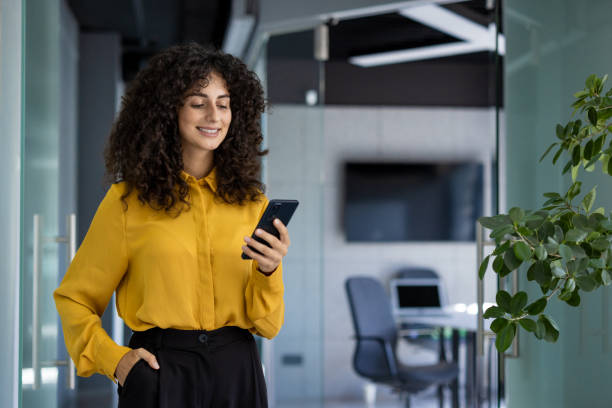 istock Smiling hispanic businesswoman using smartphone in modern office while wearing a yellow blouse, conveying confidence and connection 2180573087
