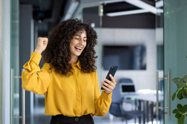 istock Excited Hispanic businesswoman celebrates success while using smartphone in modern office environment 2180574197