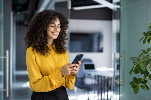 istock Hispanic businesswoman in yellow blouse smiling while using smartphone in modern office environment 2180574698