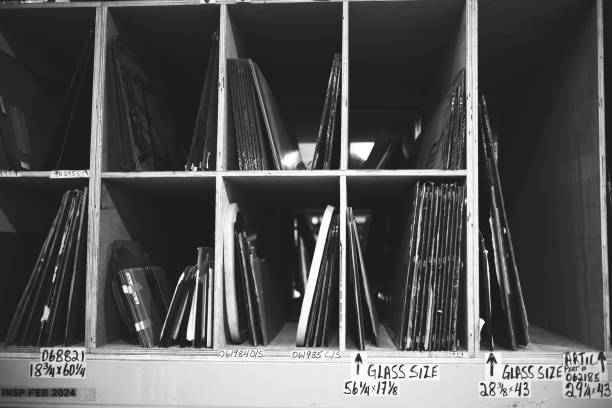 A black and white photograph of a wooden shelving unit filled with various framed artworks and glass sizes. The shelves are labeled with dimensions and identifiers, showcasing an organized display of