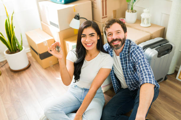 istock Happy couple displaying their new house keys amid moving boxes and a suitcase while seated on the floor 2180614308