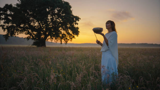 A mature Caucasian woman, clad in a white robe, plays a Tibetan singing bowl with focus, surrounded by meadow grass against a sunset backdrop.