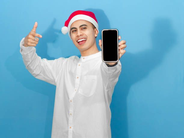 istock Excited Asian man in a white shirt and Santa hat points at a blank mobile phone screen, ready to share festive joy. 2180642245