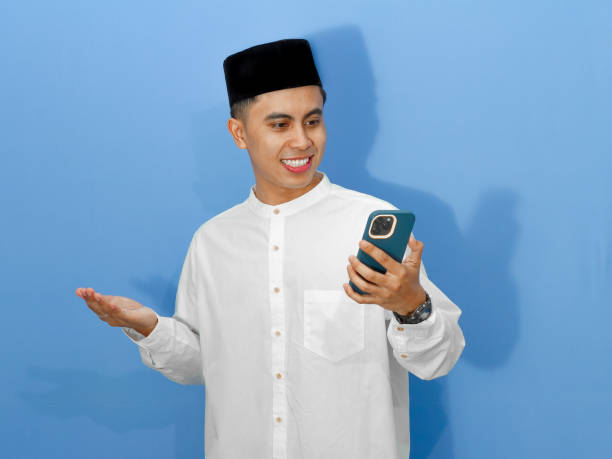 Excited Asian Muslim man using mobile phone against a blue background, ideal for Ramadan and Eid advertising lifestyle themes
