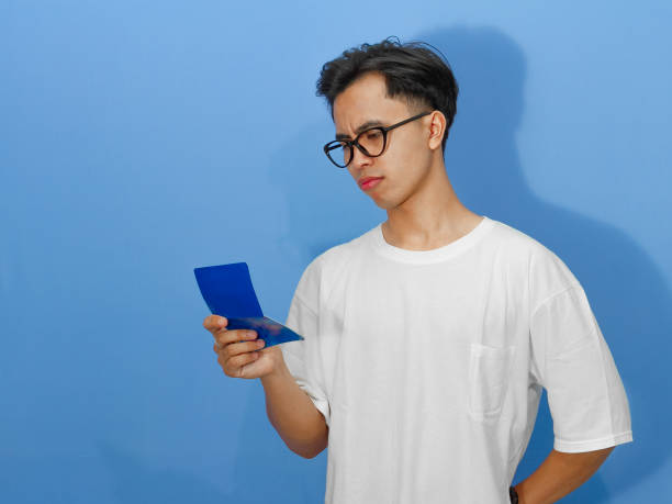 istock A worried young Asian man in a white t-shirt shows a thinking gesture while looking at his bank savings book. 2180642476