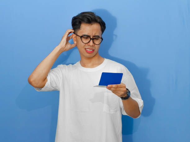 istock A worried young Asian man in a white t-shirt shows a thinking gesture while looking at his bank savings book. 2180642833