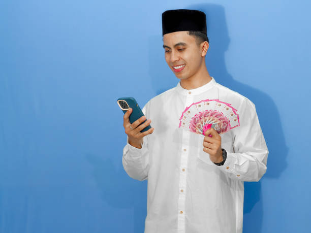 istock Smiling young Asian Muslim man holding Rupiah banknotes while looking at his mobile phone, showing a happy expression. Ideal for finance and lifestyle concepts. 2180643529