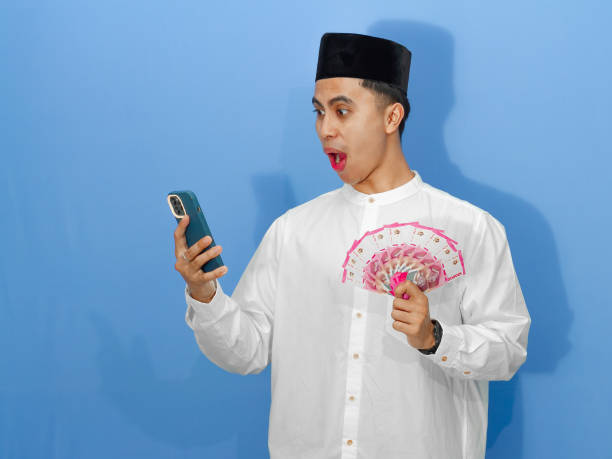 istock Happy young Asian Muslim man showing a shocked expression while using a mobile phone and holding Indonesian rupiah banknotes. 2180644302