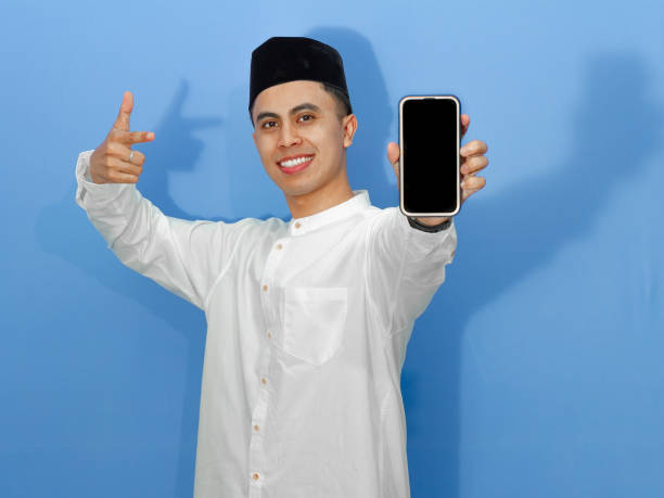 istock Excited young Asian Muslim man joyfully points at a blank mobile phone screen, showcasing potential for promotions or advertisements. 2180646834