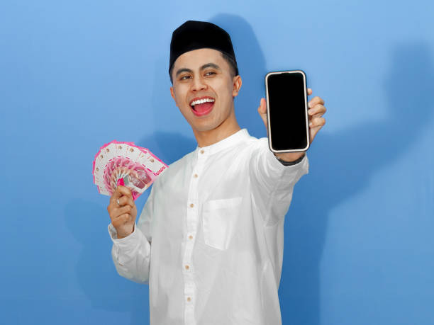 istock A happy young Asian Muslim man excitedly shows a blank mobile phone screen while holding rupiah banknotes. 2180648865
