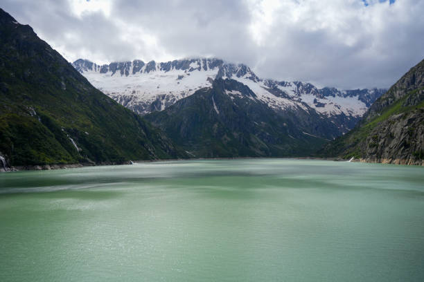 A serene turquoise alpine lake surrounded by towering snow-capped mountains and steep green slopes, creating a stunning and peaceful natural vista under a cloudy sky.