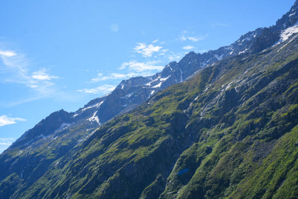 A stunning view of steep alpine slopes, covered with lush greenery and patches of snow, set against a clear blue sky with wispy clouds.