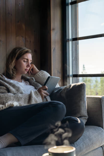 istock Woman Reading a Book With an E-Reader and Warm Drink in Sunlight Cozy Indoors 2180705897