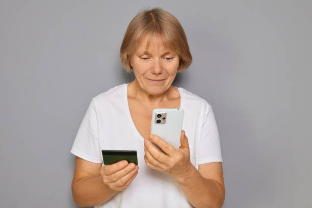 istock Senior woman smiling while paying online shopping transaction using credit card entering data on banking application with cellphone in hands standing isolated over gray background 2180715843