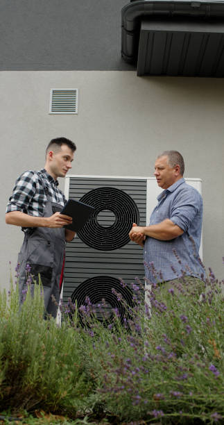 A technician discusses heat pump functionality with a homeowner in front of the unit, using a tablet for reference.