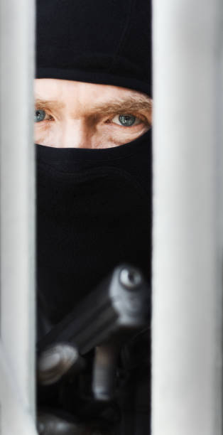 Gunman masked with balaclava looks threatening and dangerous Man holding a handgun, peering through an object while wearing a ski mask and looking like a dangerous criminal. person holding frame over face stock pictures, royalty-free photos & images