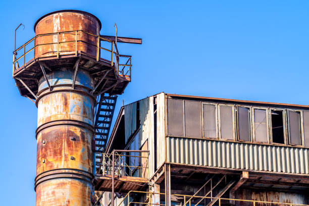 Old, rusty industrial structure with a weathered water tower against a blue sky, showing signs of abandonment with broken windows and peeling paint
