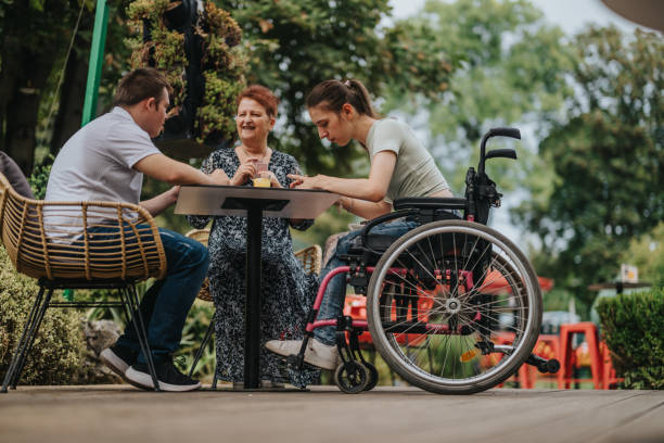 istock Three people, including one using a wheelchair, are bonding at an outdoor table, highlighting friendship and inclusivity in a lush, green setting. 2180754940