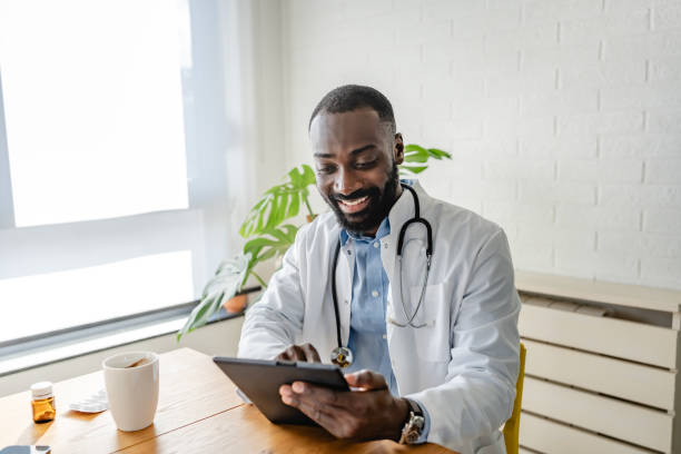 istock Smiling African-American doctor using a tablet for a virtual consultation, dressed in a white coat, with a coffee cup on the desk in a bright, modern office. 2180765266