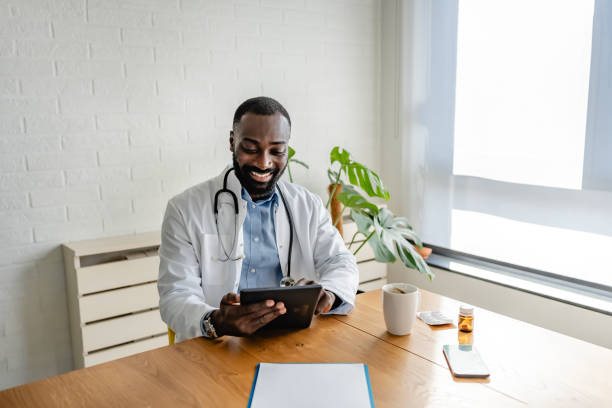 istock Smiling African-American doctor using a tablet for a virtual consultation, dressed in a white coat, with a coffee cup on the desk in a bright, modern office. 2180765276
