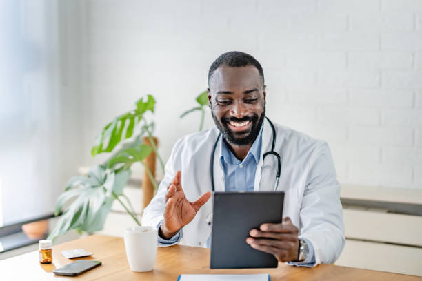 istock Smiling African-American doctor using a tablet for a virtual consultation, dressed in a white coat, with a coffee cup on the desk in a bright, modern office. 2180765363