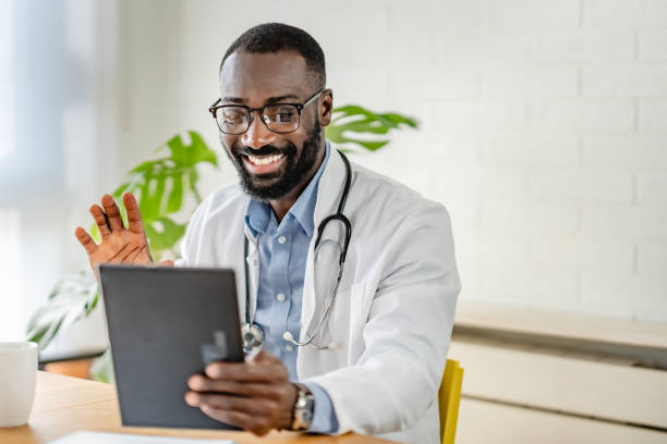 istock Smiling African-American doctor using a tablet for a virtual consultation, dressed in a white coat, with a coffee cup on the desk in a bright, modern office. 2180765581