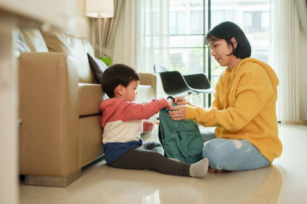 istock Asian Mother helps her little boy pack a green backpack while sitting on the floor in a living room. prepare for an outdoor activity or school. 2180770463