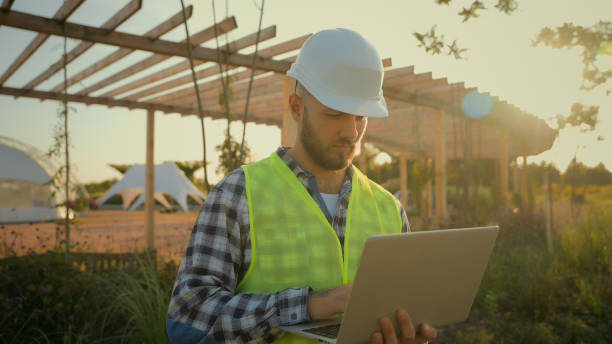 istock Thoughtful Caucasian male man experienced worker inspector helmet hardhat guy engineer technician typing laptop computer entering data internet pc analyzing project working blueprint construction park 2180774986