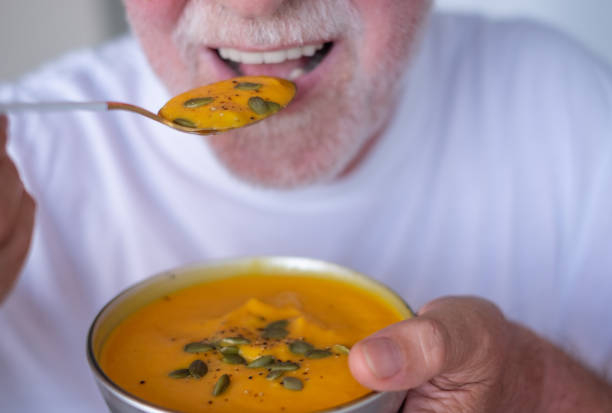 istock Cozy autumn dinner. Male hands holding a bowl with pumpkin cream soup while bringing a spoon with pumpkin cream decorated with seeds to his mouth. Healthy eating concept 2180777236