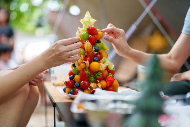 A beautifully decorated Christmas fruit tree sits on a camping table, adorned with vibrant, multicolored fresh fruits like cherry tomatoes, grapes, and berries.