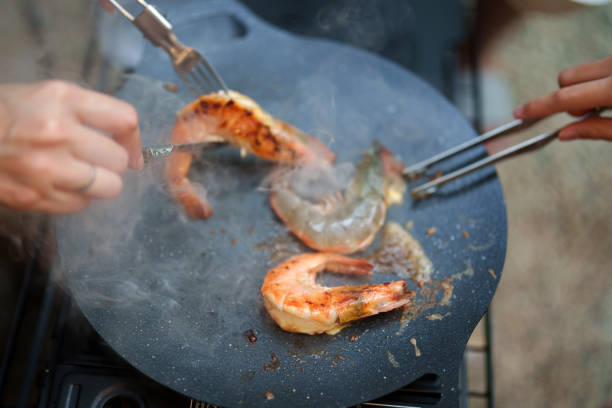 Two people are using tongs and a fork to handle the prawns, which are sizzling on the griddle and releasing steam, indicating high heat.