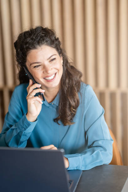 istock A cheerful woman wearing a blue blouse is sitting at a table, talking on her phone while smiling. She has a laptop in front of her, appearing to be in a work setting 2180784542