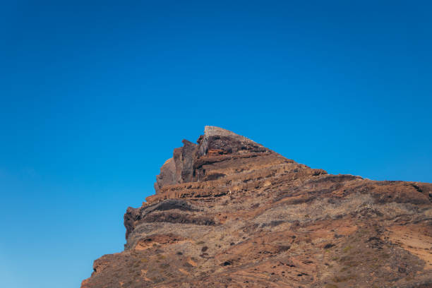 A close-up shot of a rocky mountain peak against a bright blue sky. The mountain is made up of layered rock formations, and the sky is clear and cloudless. The image is perfect for projects related to nature, landscape, mountains, rock formations, and the outdoors.