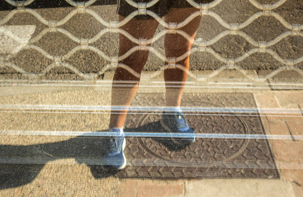 Women's legs in sportswear reflected in the glass of a shop window closed by an iron grating with a lozenge pattern. Manhole on the ground.