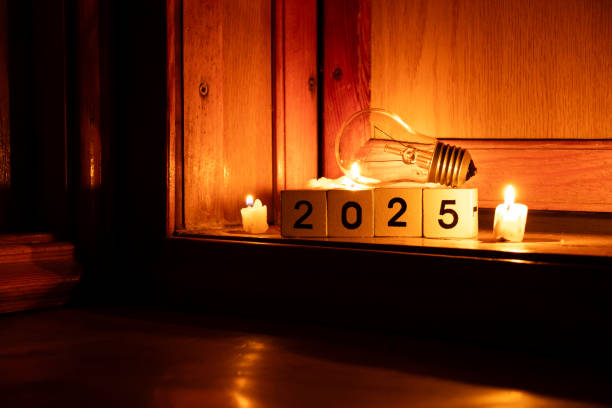2025 written on cubes and a light bulb at home on the floor with candles in the dark, new year without light stock photo