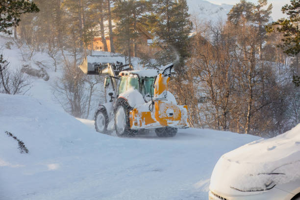 Snow plow tractor cleaning snow with a large rear-mounted snowblower, shooting snow to the side. Mountain area with snow-covered road. Effective snow removal, rural road access.