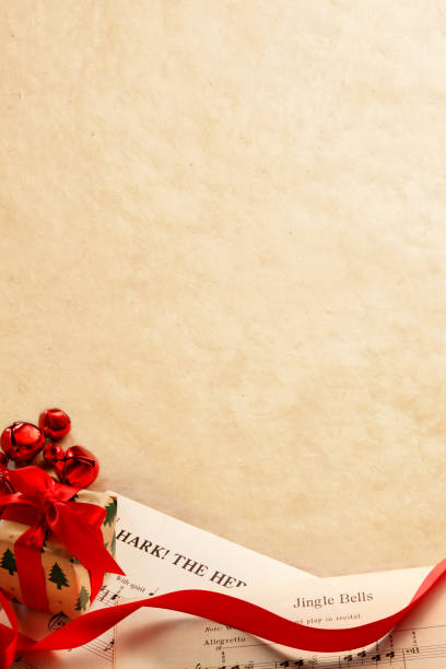 A small Christmas present is surrounded by a curled red ribbon and several red bells of differing sizes as its rests on top of the sheet music from several classic Christmas songs including Jingle Bells and Hark The Herald Angels Sing, songs that are in the public domain.