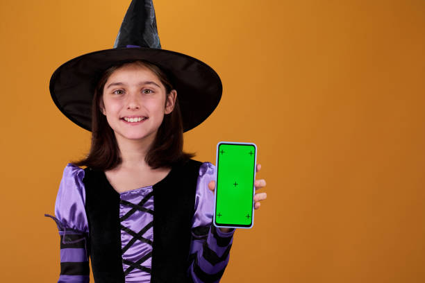 Happy cute girl in costume of Halloween witch showing you smartphone with chromakey screen with sale advert