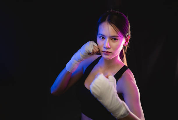 A woman is boxing in a dark room. She is wearing gloves and a black top. She is in the middle of a punch